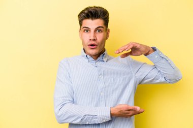 Sarı arka planda izole edilmiş genç beyaz adam iki eliyle bir şey tutuyor, ürün sunumu..