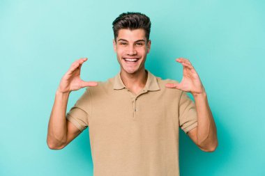 Kafkasyalı genç adam mavi arka planda izole edilmiş. Elinde avuç içi olan bir şey var ve kameraya sunuyor..