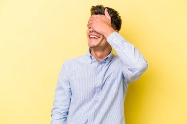 Sarı arka planda izole edilmiş genç beyaz adam elleriyle gözlerini kapatıyor, geniş bir gülümsemeyle bir sürpriz bekliyor..