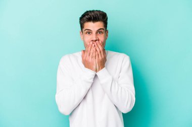 Mavi arka planda izole edilmiş genç beyaz adam şok olmuş, ağzı ellerle kaplanmış, yeni bir şey keşfetmeye hevesli..