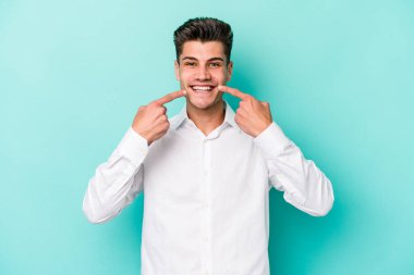 Mavi arka planda izole edilmiş genç beyaz adam gülümsüyor, parmaklarını ağıza doğrultuyor..
