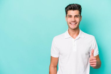 Mavi arka planda izole edilmiş genç beyaz adam gülümsüyor ve başparmağını kaldırıyor.