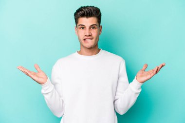 Kafkasyalı genç adam mavi arka planda izole edilmiş. Kafası karışmış ve omuz silkiyor. Kopya alanını tutmak için..