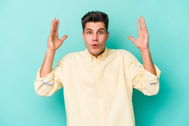 Mavi arka planda izole edilmiş genç beyaz bir adam zaferi ya da başarıyı kutluyor, şaşırıyor ve şok oluyor..