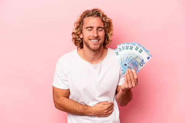 Young caucasian man holding banknotes isolated on pink background laughing and having fun.