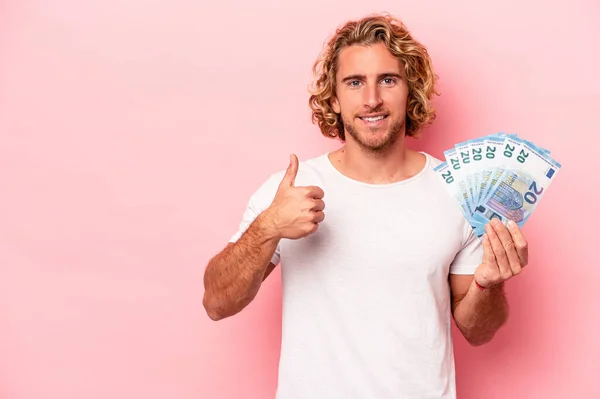 Young caucasian man holding banknotes isolated on pink background smiling and raising thumb up