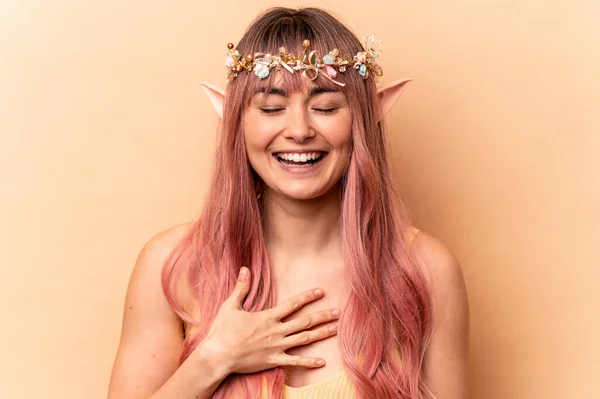 Young elf woman with pink hair isolated on beige background laughs out loudly keeping hand on chest.