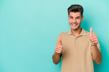 Mavi arka planda izole edilmiş genç beyaz adam gülümsüyor ve başparmağını kaldırıyor.