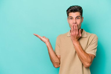 Mavi arka planda izole edilmiş genç beyaz adam, avuç içi fotokopi alanını elinde tutmaktan etkilenmiş..