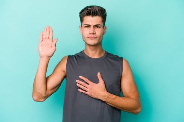 Kafkasyalı genç adam mavi arka planda izole edilmiş yemin ediyor, elini göğsüne koyuyor..