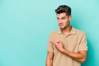 Mavi arka planda izole edilmiş genç beyaz adam gülümsüyor ve boş alanda bir şeyler gösteriyor..