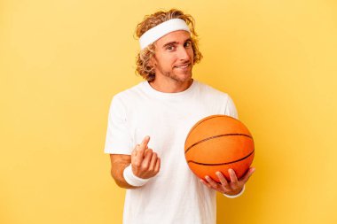 Young basketball player caucasian man isolated on yellow background pointing with finger at you as if inviting come closer.