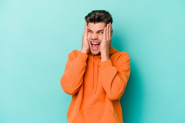 Kafkasyalı genç adam mavi arka planda izole edilmiş kulakları kulaklarıyla kapatıyor çok fazla ses duymamaya çalışıyor..