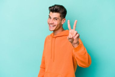 Kafkasyalı genç adam mavi arka planda izole edilmiş neşeli ve kaygısız parmaklarıyla barış sembolü gösteriyor..