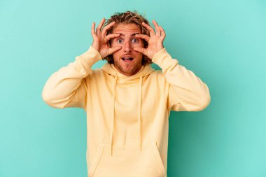 Mavi arka planda izole edilmiş genç beyaz adam bir başarı fırsatı bulmak için gözlerini açık tutuyor..