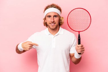 Badminton oynayan beyaz bir genç, pembe arka planda izole edilmiş, gömlek fotokopi alanını işaret ediyor, gururlu ve kendinden emin.