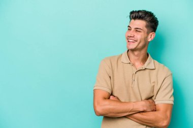 Mavi arka planda izole edilmiş genç beyaz adam kollarını kavuşturarak gülümsüyor..