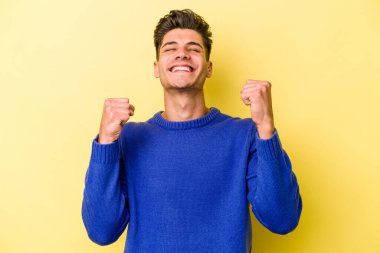 Sarı arka planda izole edilmiş genç beyaz adam zaferi, tutkuyu ve coşkuyu kutluyor, mutlu bir ifade..