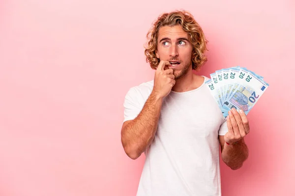 Young caucasian man holding banknotes isolated on pink background relaxed thinking about something looking at a copy space.