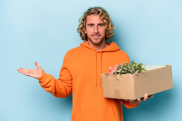 Young caucasian man making a move while picking up a box full of things isolated on blue background showing a copy space on a palm and holding another hand on waist.