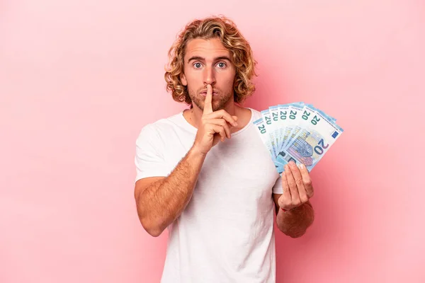 Young caucasian man holding banknotes isolated on pink background keeping a secret or asking for silence.