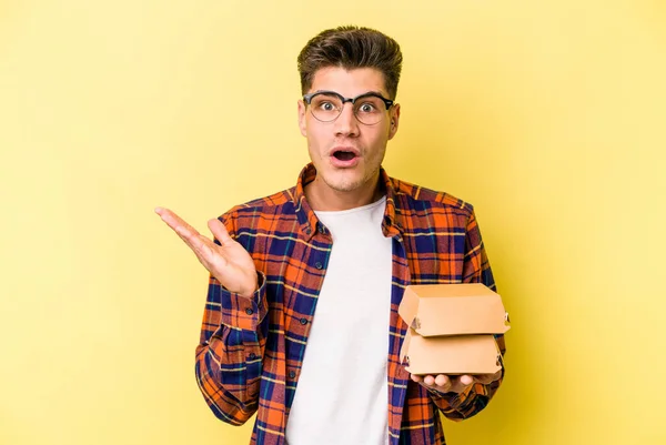Young Caucasian Man Holding Burger Isolated Yellow Background Surprised Shocked — Stockfoto