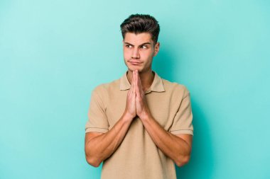 Kafkasyalı genç adam mavi arka planda izole edilmiş dua ediyor, bağlılık gösteriyor, dindar biri ilahi ilham arıyor..