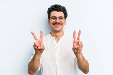 Mavi arka planda izole edilmiş genç beyaz adam zafer işareti gösteriyor ve gülümsüyor..