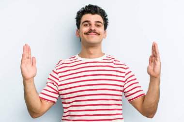 Kafkasyalı genç adam mavi arka planda izole edilmiş. Elinde işaret parmaklarıyla küçük bir şey tutuyor, gülümsüyor ve kendinden emin..