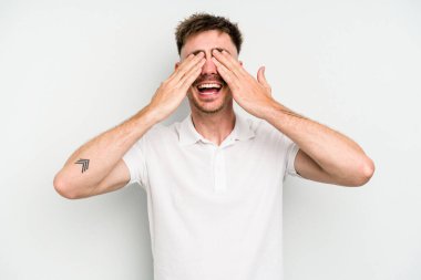 Beyaz arka planda soyutlanmış beyaz tenli genç bir adam elleriyle gözlerini kapatıyor, geniş bir gülümsemeyle bir sürpriz bekliyor..