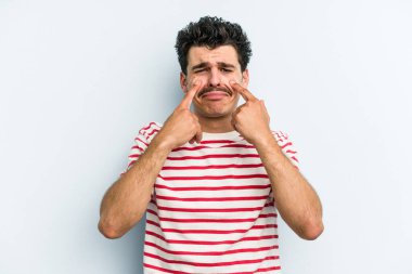 Kafkasyalı genç adam mavi arka planda izole edilmiş, ağlıyor, bir şeyden memnun değil, acı ve kafa karışıklığı konsepti..