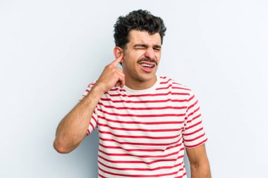 Genç beyaz adam mavi arka planda izole edilmiş parmaklarıyla kulaklarını kapatıyor, gürültülü bir ortam tarafından stresli ve umutsuz..