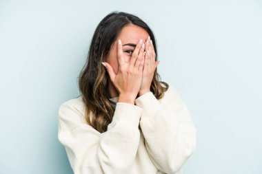 Mavi arka planda izole edilmiş genç beyaz kadın kameraya parmaklarıyla göz kırpıyor, yüzünü kapatmaktan utanıyor..
