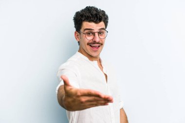 Mavi arka planda izole edilmiş genç beyaz adam selamlama jesti olarak kameraya elini uzatıyor..