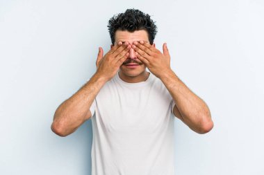 Mavi arka planda izole edilmiş genç beyaz adam elleriyle gözlerini kapatmaktan korkuyor..