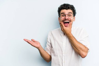 Mavi arka planda izole edilmiş genç beyaz bir adam avucunun içinde fotokopi alanı tutuyor, el üstünde tutuyor. Şaşırmış ve mutlu..