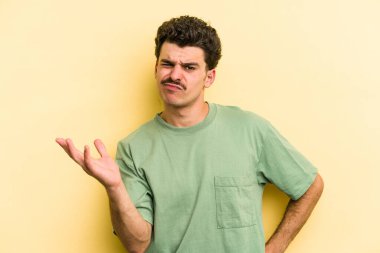 Sarı arka planda izole edilmiş genç beyaz adam sorgulama sırasında omuz silkiyor ve kuşkulanıyor..