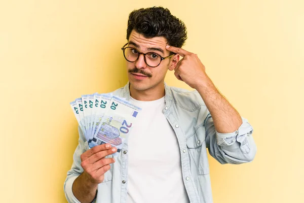 Young caucasian man holding banknotes isolated on yellow background pointing temple with finger, thinking, focused on a task.