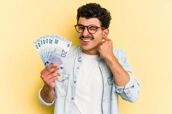 Young caucasian man holding banknotes isolated on yellow background covering ears with hands.