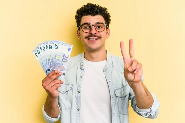 Young caucasian man holding banknotes isolated on yellow background showing number two with fingers.