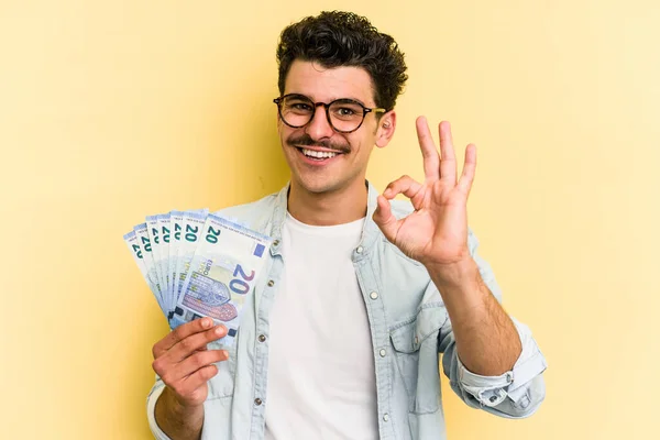 Young caucasian man holding banknotes isolated on yellow background cheerful and confident showing ok gesture.