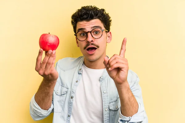 Young caucasian man holding an apple isolated on yellow background having an idea, inspiration concept.