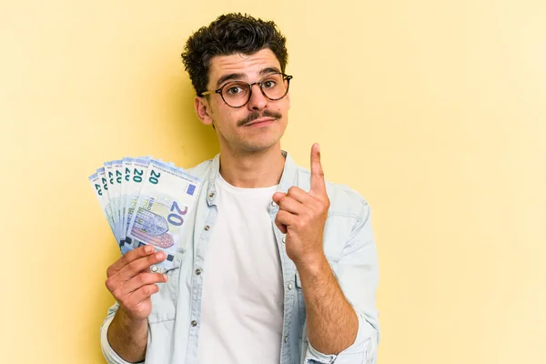 Young caucasian man holding banknotes isolated on yellow background showing number one with finger.