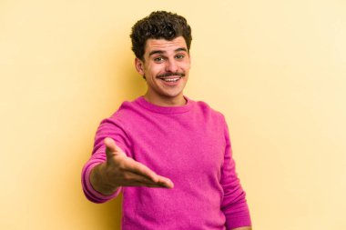 Sarı arka planda izole edilmiş genç beyaz adam selamlama jesti olarak kameraya elini uzatıyor..