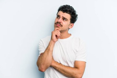 Kafkasyalı genç adam mavi arka planda izole edilmiş düşünüyor ve bakıyor, yansıtıyor, düşünüyor, bir fantezi kuruyor..