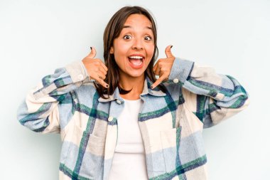 Genç İspanyol kadın mavi arka planda yalnız mutlu ve kendinden emin gülümsüyor, elleriyle çenesine dokunuyor..