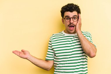 Sarı arka planda izole edilmiş genç beyaz adam, avuç içi fotokopi alanını elinde tutmaktan etkilendi..