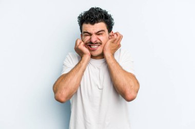 Mavi arka planda izole edilmiş genç beyaz adam kulaklarını elleriyle kapatıyor..