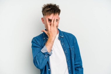 Beyaz arka planda izole edilmiş genç beyaz bir adam kameraya parmaklarıyla göz kırpıyor, yüzünü kapatmaktan utanıyor..