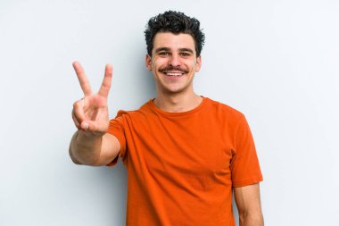 Mavi arka planda izole edilmiş genç beyaz adam zafer işareti gösteriyor ve gülümsüyor..